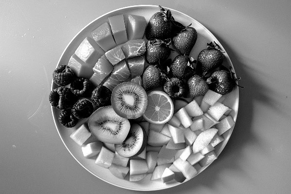 A plate with an assortment of fresh fruits, including sliced kiwi, raspberries, strawberries, orange slices, and cubes of melon, arranged neatly in sections.